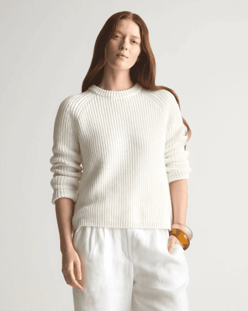 A woman with long red hair wears a textured white sweater and loose white pants, accessorized with chunky bracelets on her left wrist, standing against a plain light background, by McGee & Co.